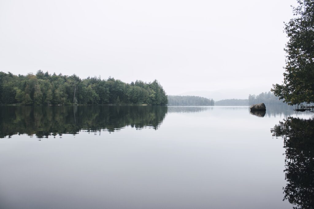 Serene lake with still waters reflecting a forested landscape and overcast sky.